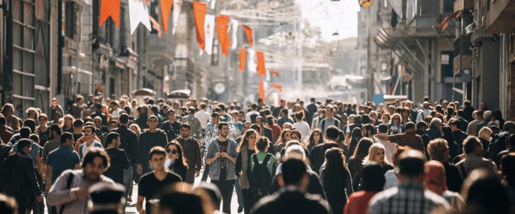 Folla di persone che passeggiano lungo una via cittadina affollata, con bandiere appese sopra la strada e negozi ai lati.